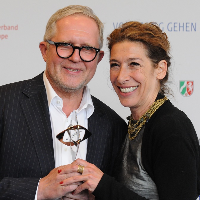 Harald Krassnitzer und Adele Neuhauser stehen lächelnd vor einer Logowand und halten gemeinsam eine Statue des Grimme Preises in den Händen.