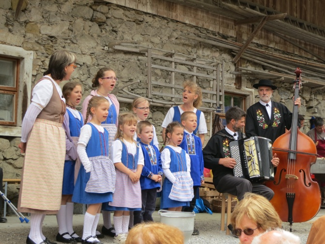 Eine Gruppe von Menschen in Tracht steht zusammen und jodelt. 2 Männer spielen Akkordeon und Kontrabass.
