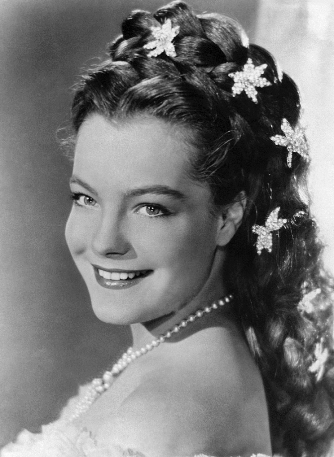 Schwarz-weiß-Foto von Romy Schneider mit aufwendiger Frisur und sternförmigem Haarschmuck. Sie lächelt im Seitenprofil in die Kamera.