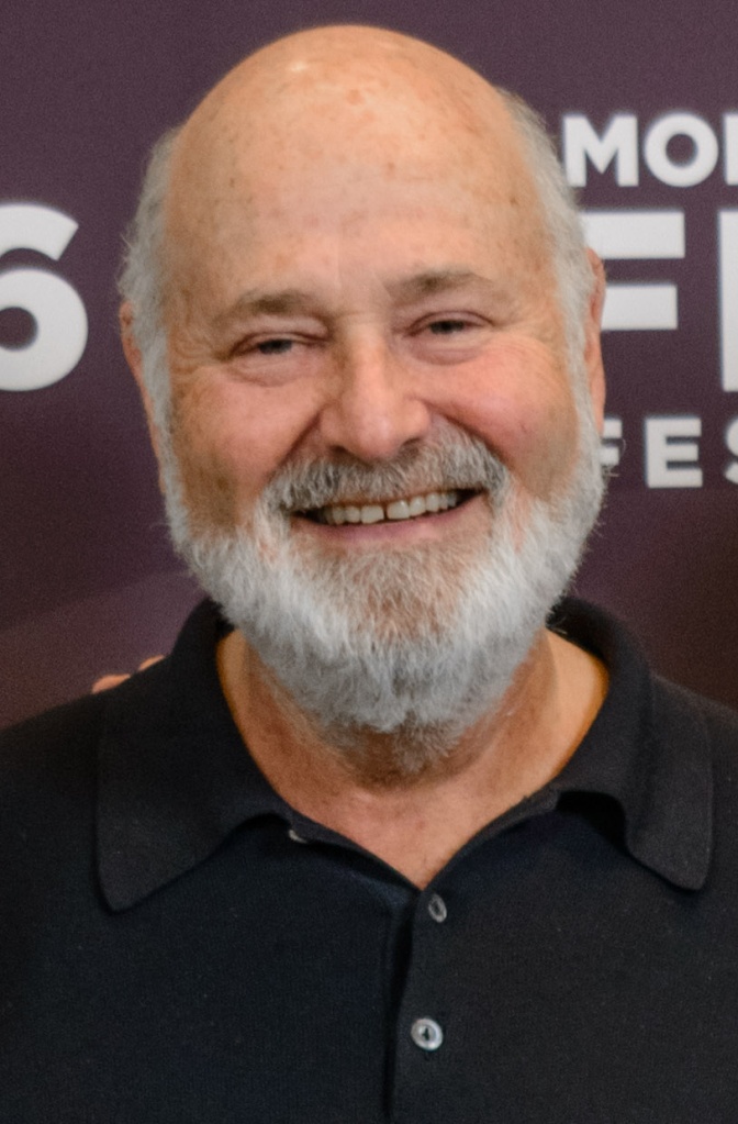 Rob Reiner mit Glatze und weißem Vollbart. Er schaut lächelnd direkt in die Kamera und steht vor einer Logowand.