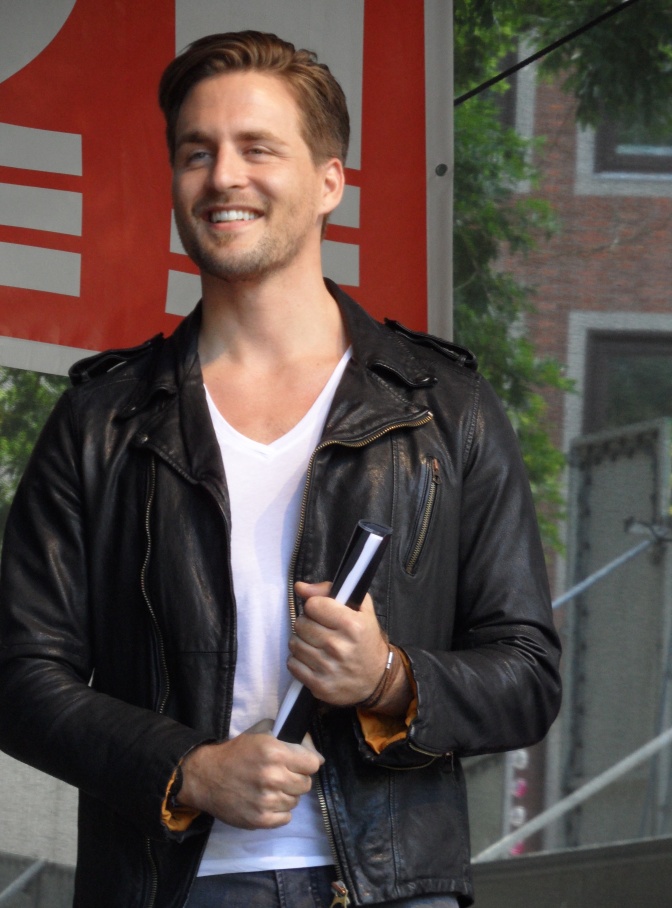 Alexander Klaws steht in Lederjacke lächelnd auf einer Bühne. Er hält eine Rolle aus zusammengerolltem Papier in beiden Händen.