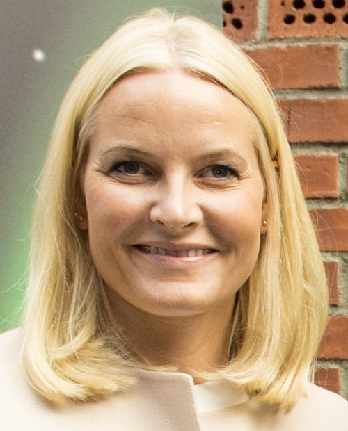 Mette-Marit mit schulterlangen, glatten blonden Haaren. Sie steht vor einer Ziegelwand und lächelt in die Kamera.