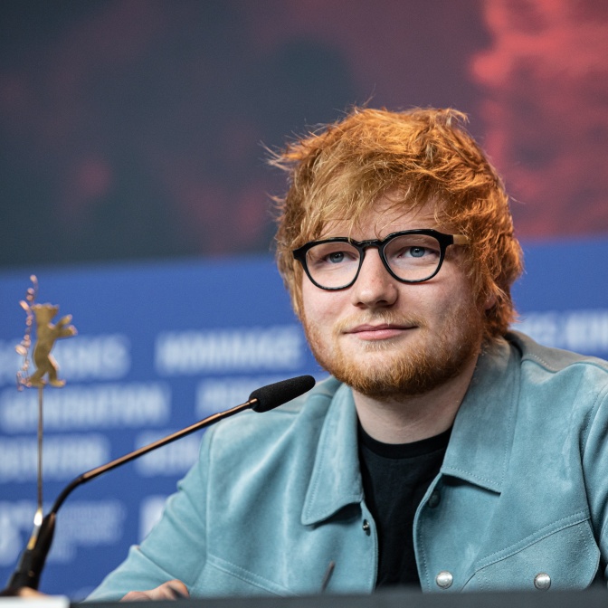 Ed Sheeran mit Brille und Vollbart. Er sitzt vor einem Mikrofon und schaut direkt in die Kamera.