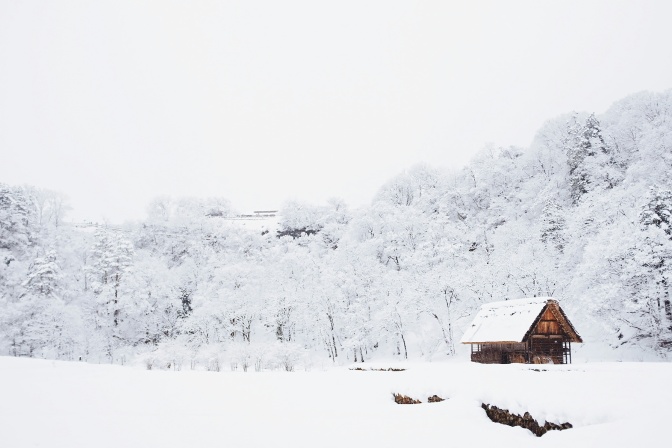 Schneebedeckter Wald, im Vordergrund eine kleine Hütte auf einer Lichtung. Das Dach der Hütte ist auch mit Schnee bedeckt, der Himmel ist weiß.