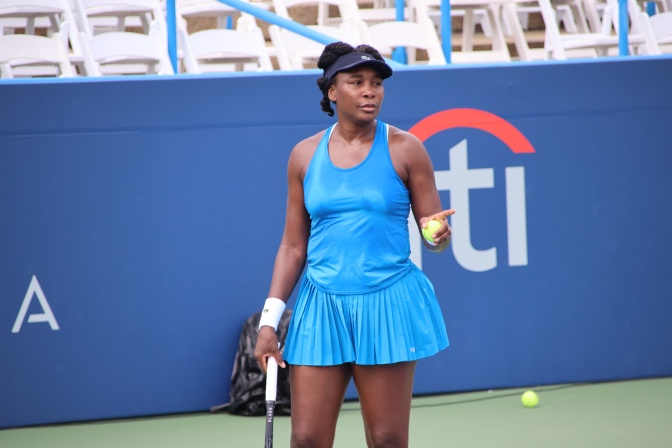 Venus Williams in einem blauen Tennisdress auf dem Tennisplatz. Sie ist schwarz und trägt ihre Haare zu Zöpfen gezwirbelt.
