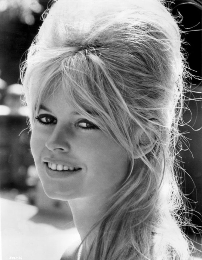 Schwarz-weiß-Foto einer jungen Brigitte Bardot mit einer hoch aufgetürmten Hochsteckfrisur. Sie schaut lächelnd in die Kamera.