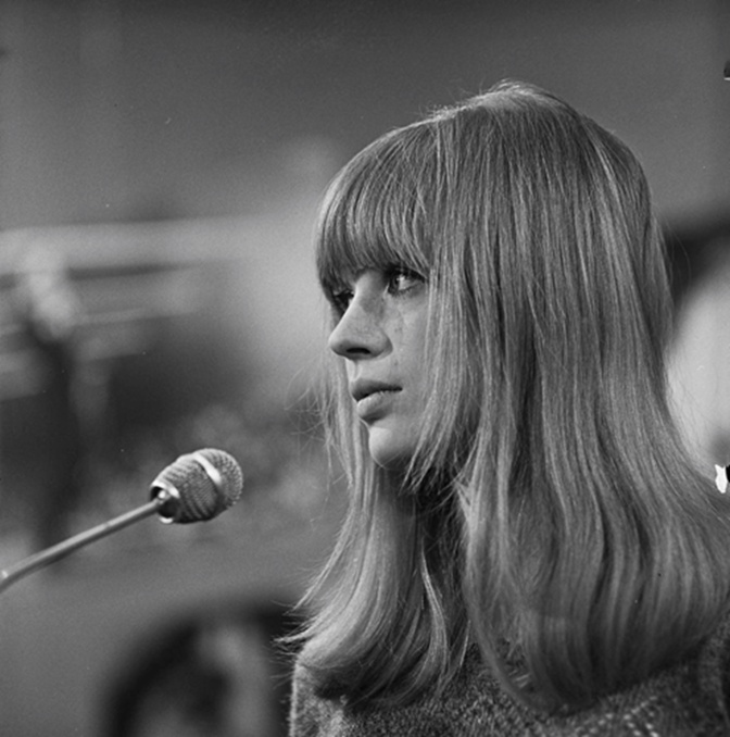 Schwarz-weiß-Foto einer jungen Marianne Faithfull am Mikrofon. Sie hat lange glatte Haare und einen voluminösen Pony. Sie ist im Seitenprofil zu sehen.