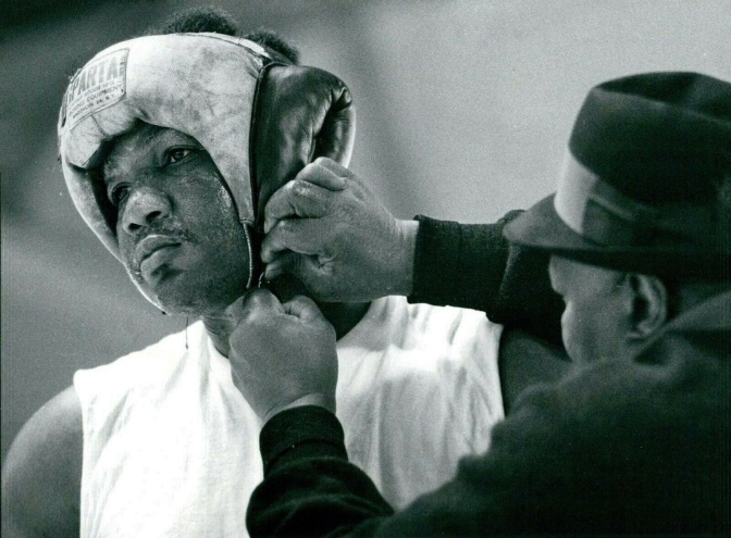 Schwarz-weiß-Foto eines jungen George Foreman im Boxring. Jemand schließt seinen Kopfschutz aus Leder.