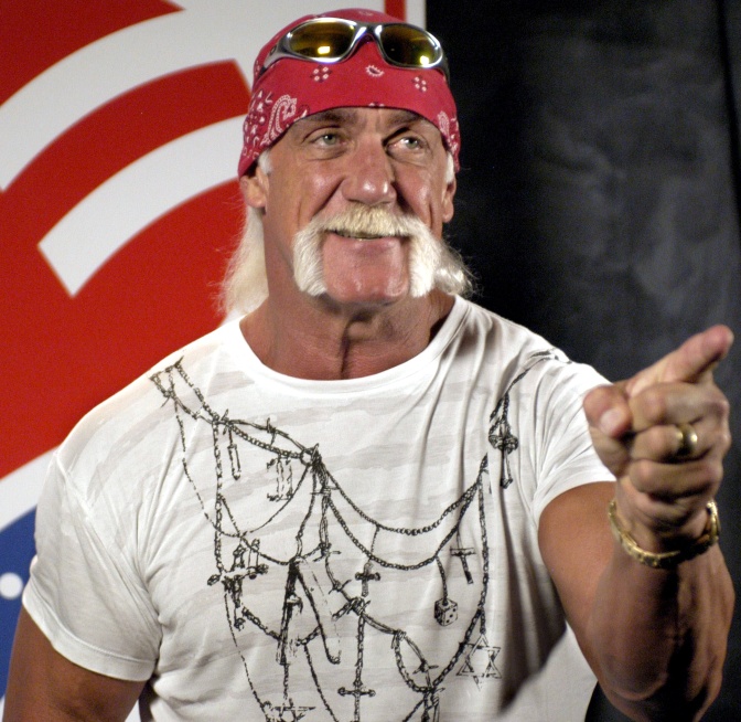 Hulk Hogan mit einem Bandana und einer Sonnenbrille auf der Stirn. Er steht vor einer amerikanischen Flagge und zeigt mit dem Finger in Richtung der Kamera.