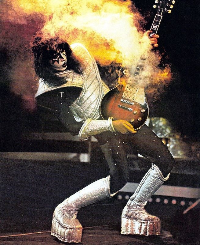 Ace Frehley mit weiß-schwarz geschminktem Gesicht in einem Bühnenkostüm mit silbernen Plateaustiefeln. Im Hintergrund Flammen. Es spielt Gitarre.