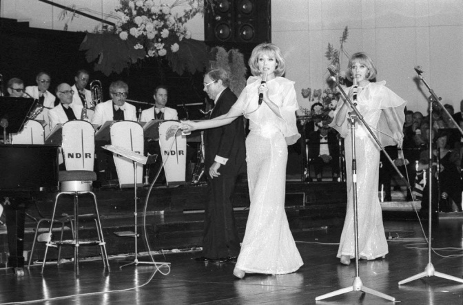 Schwarz-weiß-Fotos der Kessler-Zwillinge. Sie stehen in festlichen Kleidern nebeneinander auf der Bühne und singen.