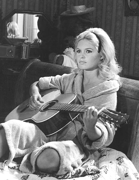Schwarz-weiß-Foto einer jungen Brigitte Bardot. Sie sitzt auf einem Sofa und spielt eine akustische Gitarre. Sie schaut mit starrem Blick an der Kamera vorbei.