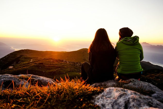 2 Frauen sitzen nebeneinander im Freien. Sie sind von hinten fotografiert und sitzen im Gegenlicht. Sie tragen Mützen und Jacken.