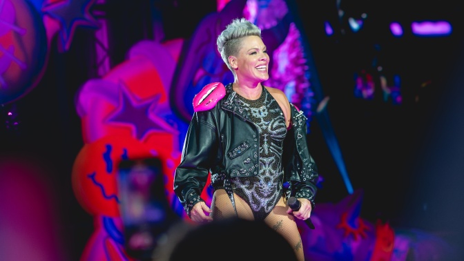 Pink auf der Bühne in einem Body mit Strass und in schwarzer Nietenlederjacke. Sie lächelt und hält ein Mikro in der Hand.