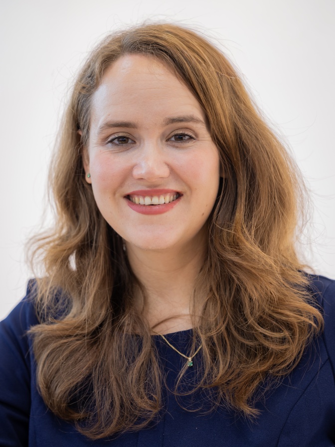 Ricarda Lang mit langen, gewellten hellbraunen Haaren und rotem Lippenstift. Sie schaut direkt in die Kamera und lächelt.