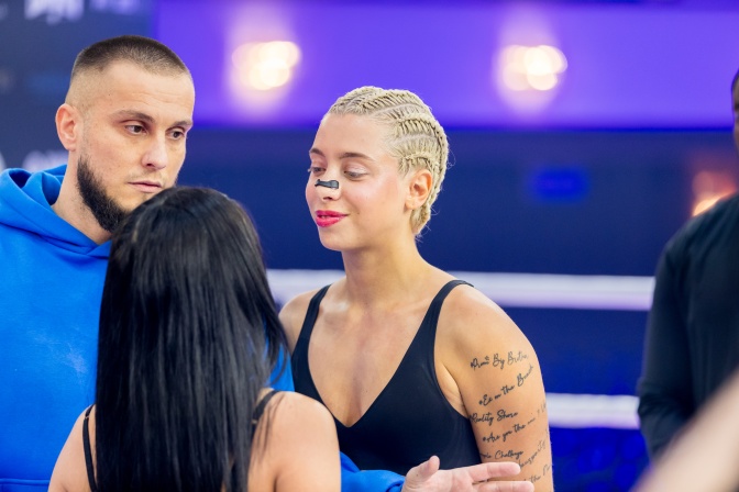 Eine junge Frau mit blondierten, zu eng am Kopf liegenden Zöpfen geflochtenen Haaren. Sie hat ein Pflaster auf der Nase kleben und wird für einen Boxkampf fit gemacht. 2 Personen stehen neben ihr und sprechen mit ihr.