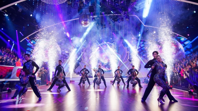 Tänzer*innen auf einer Bühne, hinter ihnen werden weiße Feuerwerksäulen gezündet.