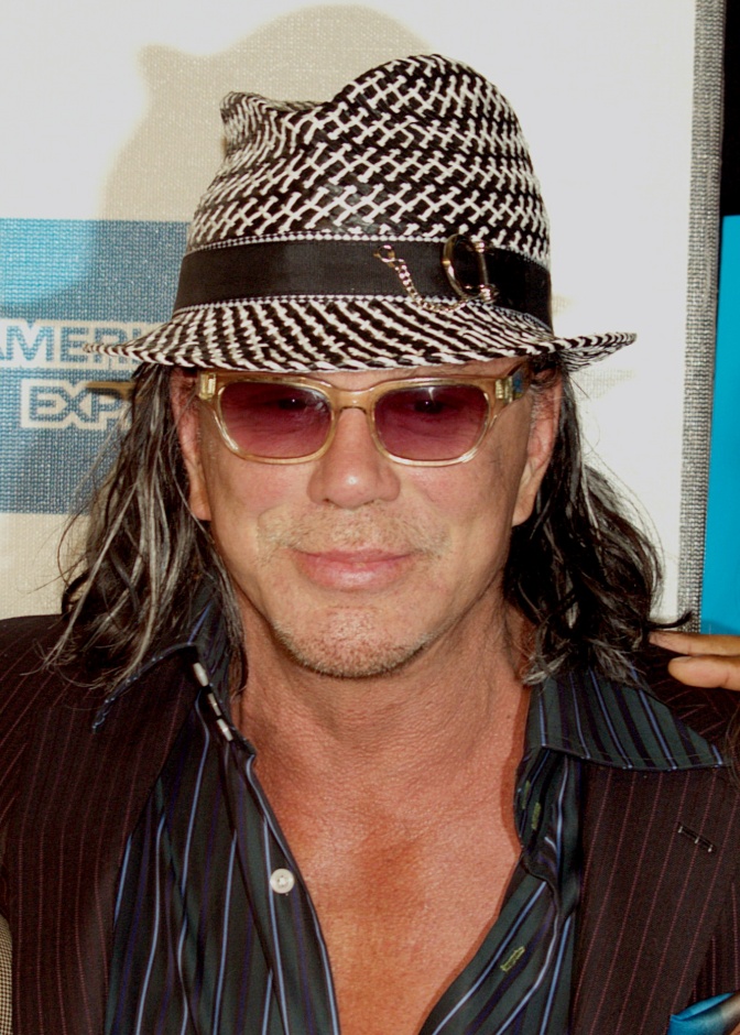 Mickey Rourke mit langen gewellten Haaren. Er trägt einen Hut und Sonnenbrille. Sein Hemd ist bis zur Brust aufgeknöpft.