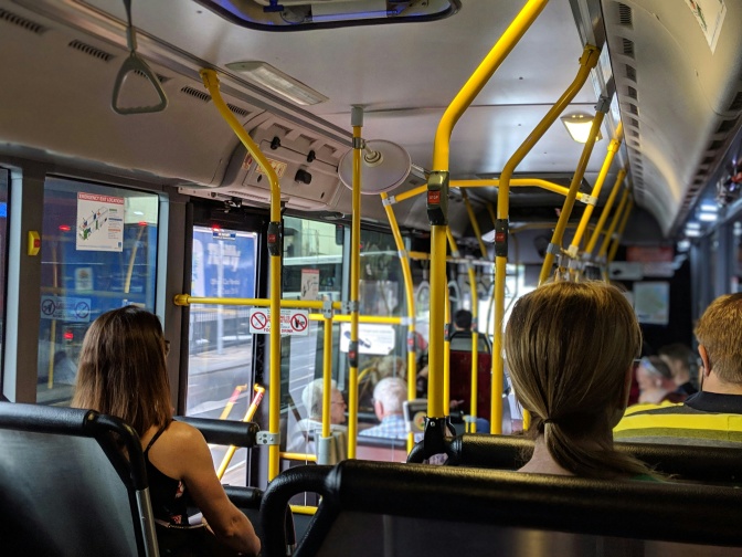 Mehrere Fahrgäste in einem Stadtbus. Die sitzenden Menschen sind nur von hinten zu sehen.