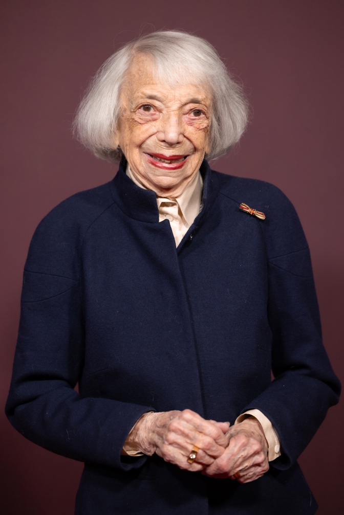 Margot Friedländer mit weißen Haaren und in einem dunkelblauen Kostüm. Sie trägt leuchtend roten Lippenstift und schaut mit einem leichten Lächeln in die Kamera.