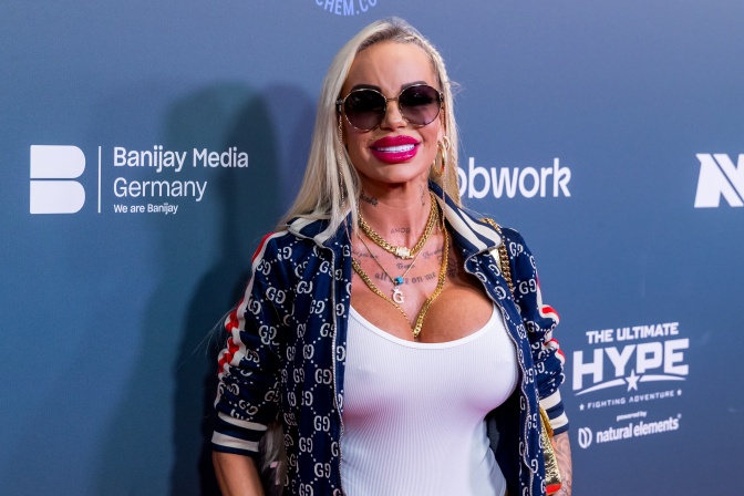 Gina-Lisa Lohfink in körperbetonter Kleidung vor einer Logowand. Sie trägt eine dunkle Sonnenbrille und lächelt in die Kamera.