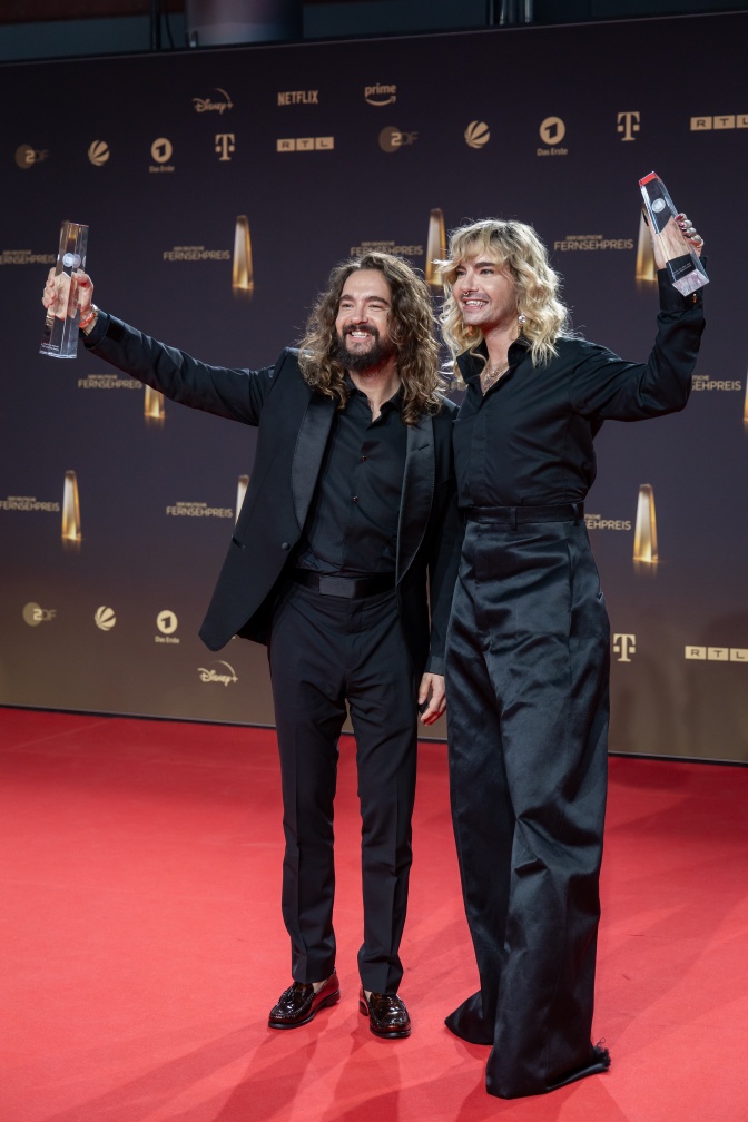 Bill und Tom Kaulitz stehen lächelnd nebeneinander vor einer Logowand. Sie halten je eine Preisstatue aus Plexiglas in der Hand.