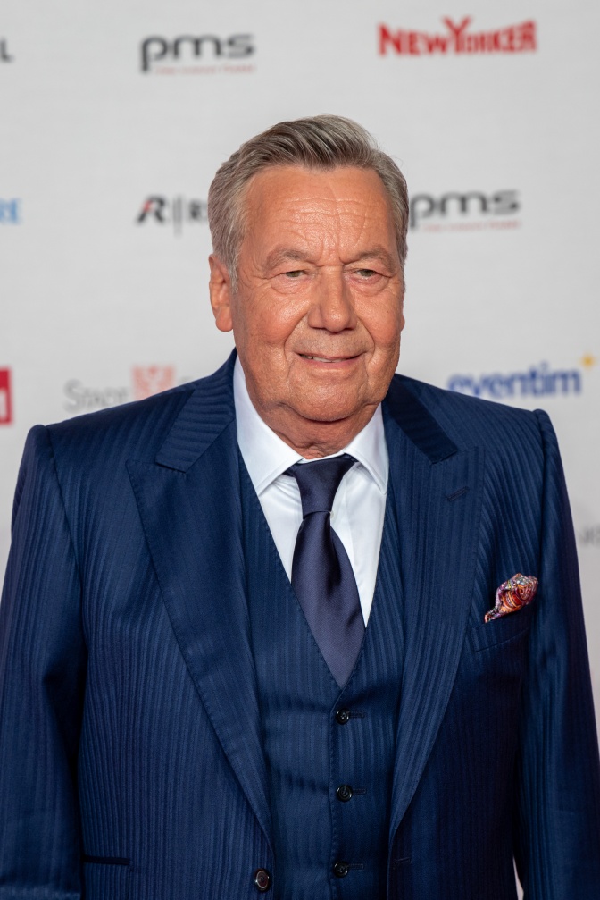 Roland Kaiser steht in einem dunkelblauen Anzug vor einer Logowand.