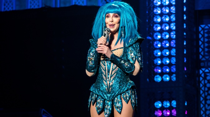 Cher auf der Bühne in einem glitzernden Bühnenkostüm in verschiedenen Blautönen und mit blauer Perücke. Sie hat ein Mikrofon in der Hand und lächelt.