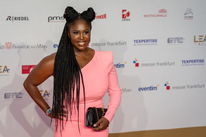 Motsi Mabuse in einem asymmetrischen rosafarbenen Kleid vor einer Logowand.