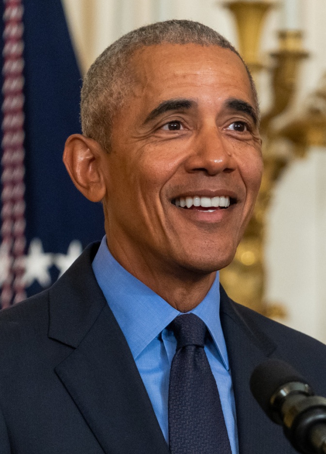 Barack Obama in blauem Hemd mit Anzug und Krawatte. Er schaut seitlich an der Kamera vorbei und lächelt. Seine Haare sind sehr kurz und weiß.