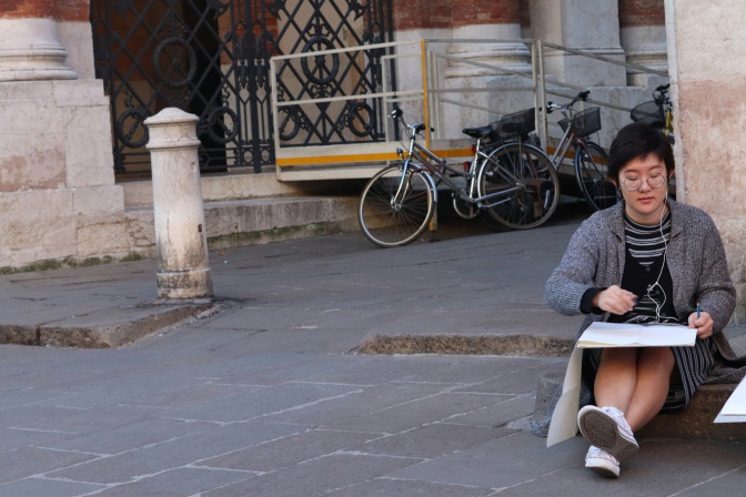 Eine junge asiatische Frau sitzt im Freien auf dem Boden und zeichnet.