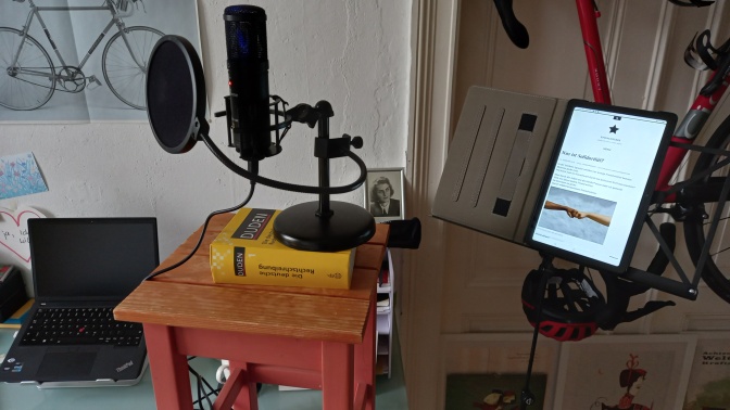 Ein Mikrofon an einem Ständer steht auf einem kleinen Tisch. Darunter liegt ein Buch, damit die Höhe passt.