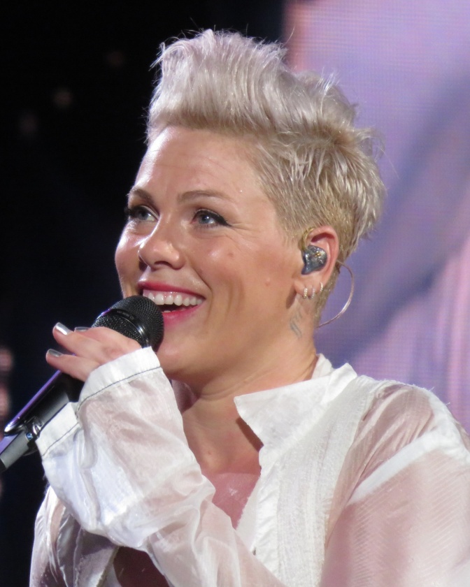 P!nk mit zur Tolle frisierten blondierten Haaren. Sie trägt eine weiße Bluse und spricht in ein Mikro.
