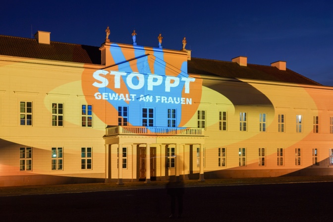 Ein großes Gebäude wird orangefarbne angeleuchtet, dazu wird der Schriftzug Stoppt Gewalt an Frauen an die Fassade projiziert.