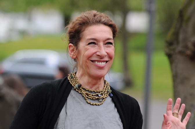 Adele Neuhauser lächelt in die Kamera und winkt. Sie hat die Haare hochgesteckt und trägt festliche Kleidung.