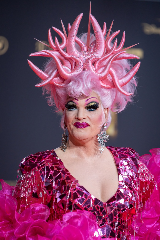 Olivia Jones  in einem aufwendigen Kostüm in Pink- und Rosatönen. Sie trägt starkes Make up und schaut direkt in die Kamera.