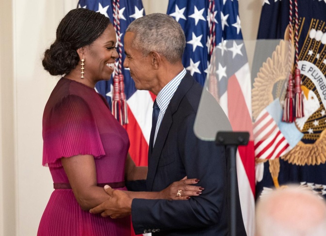 Michelle und Barack Obama in festlicher Kleidung. Beide umarmen sich und sind von der Seite zu sehen, im Hintergrund hängt eine amerikanische Flagge.