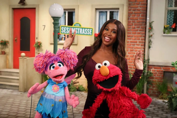 Motsi Mabuse steht neben einem Stra0enschild der Sesamstraße und lächelt. Um sie herum stehen mehrere Figuren der Sesamsraße, unter anderem Elmo.