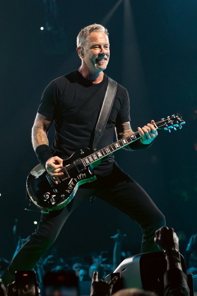 James Hetfield auf der Bühne. Er trägt schwarz und spielt E-Gitarre.