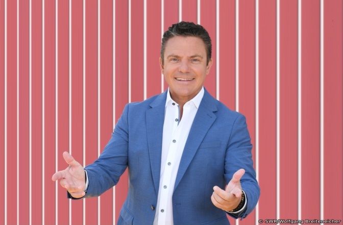 Stefan Mross mit ausgebreiteten Armen lächelnd vor einer gestreiften Tapete.