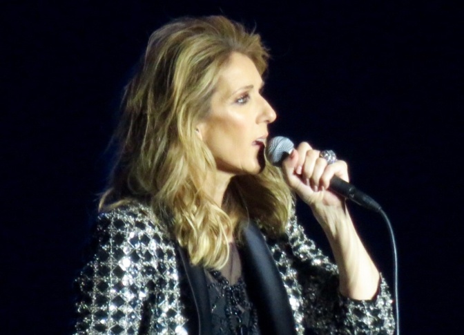 Céline Dion singt in ein Handmikro. Sie hat lange, blonde gewellte Haare und trägt ein kariertes Sakko.