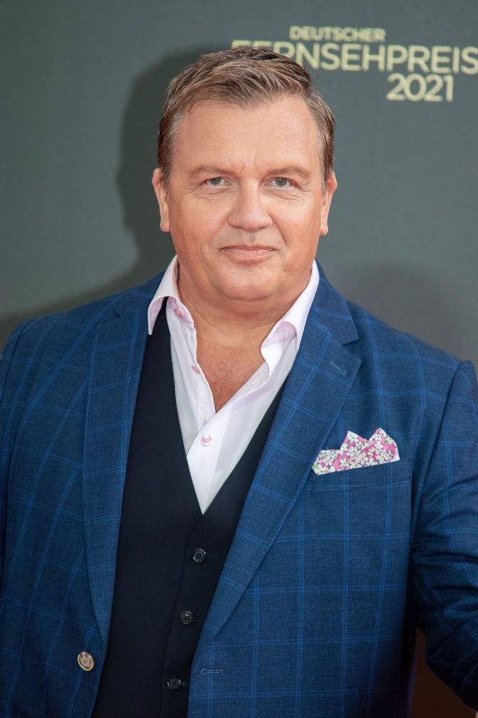 Hape Kerkeling in blauem Sakko und Hemd. Er steht mit ernstem Blick vor einer Logowand.