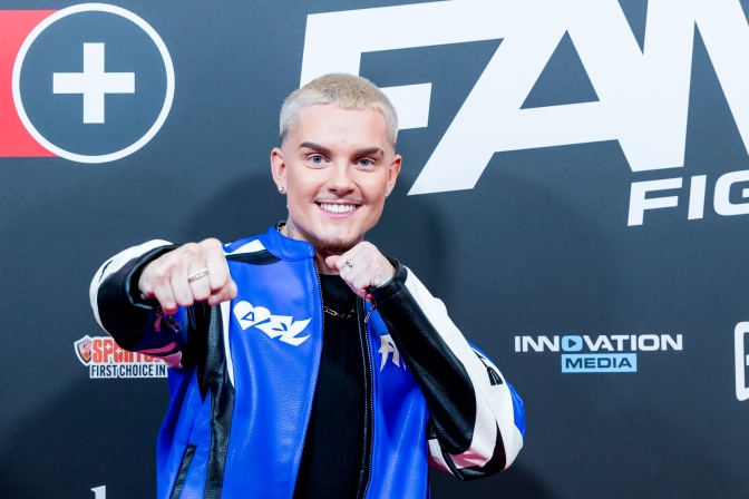 Tim Kampmann steht in einer Sportjacke vor einer Logowand und deutet einen Boxschlag an. Er lächelt in die Kamera.