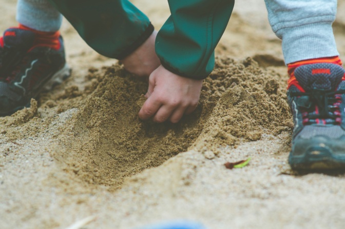Ein Kind buddelt im Sand. Man sieht nur Hände und Füße des Kindes.