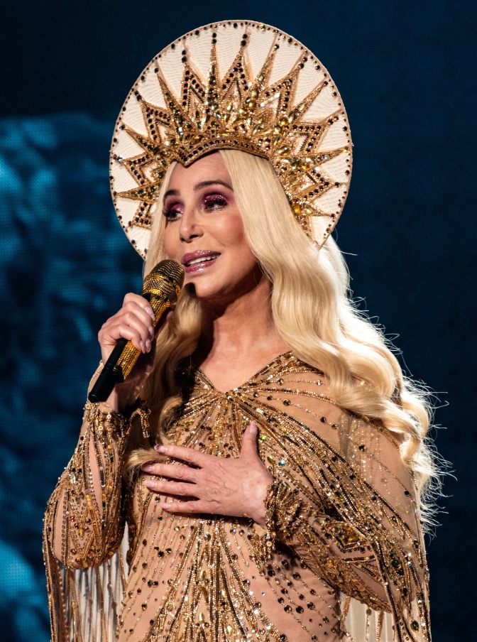 Cher in einem goldenen Bühnenkostüm mit einem Heiligenschein auf dem Kopf. Sie hat eine blonde Perücke auf und singt in ein Mikrofon.