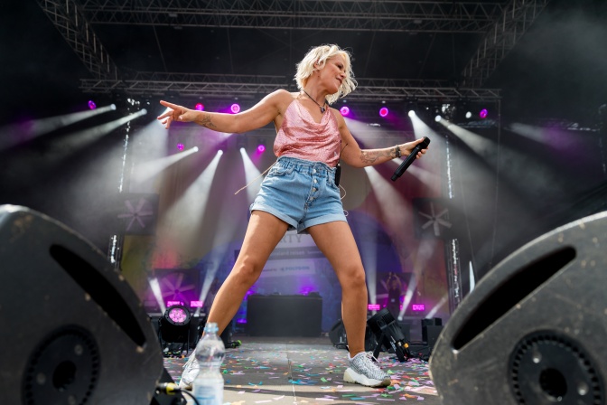Michelle in Jeansshorts auf der Bühne. Sie hat ein Mikrofon in der Hand, hinter ihr Scheinwerfer.
