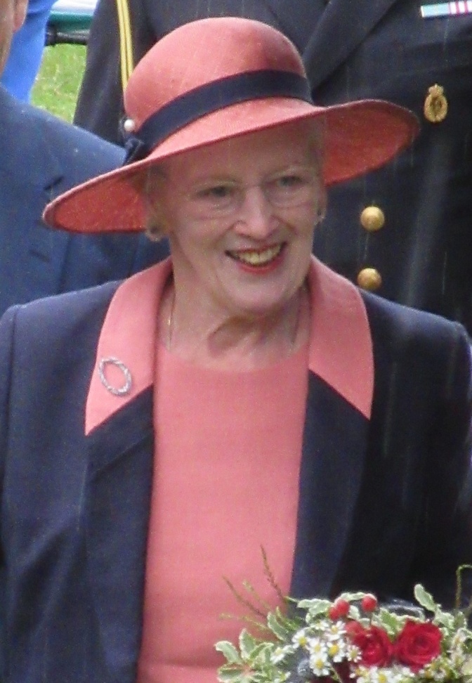 Margrethe mit einem pfirsichfarbenen Hut und passender Bluse. Darüber trägt sie eine dunkelblaue Jacke und sie hat einen Blumenstrauß in der Hand.