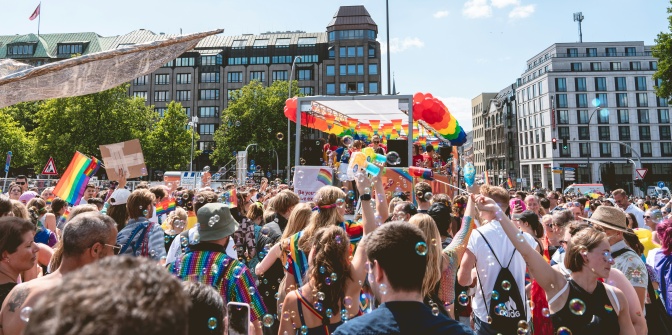 Viele Menschen bei einer Straßenparade. Viele von ihnen tragen Regenbogenflaggen. Im Hintergrund sieht man einen geschmückten LKW mit Girlanden und Regenbogen.
