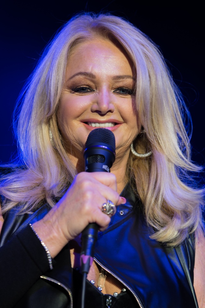 Bonnie Tyler mit blonden, gewellten Haaren. Sie hält ein Mikro in der Hand und lächelt.