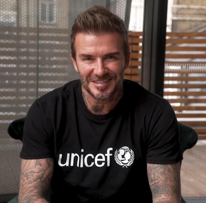 David Beckham in einem schwarzen Shirt mit Unicef-Print auf der Brust. Er trägt einen Vollbart und lächelt in die Kamera.